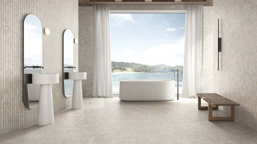 Baño con bañera exenta y suelo y paredes de porcelánico claro de aspecto piedra, frente a un ventanal